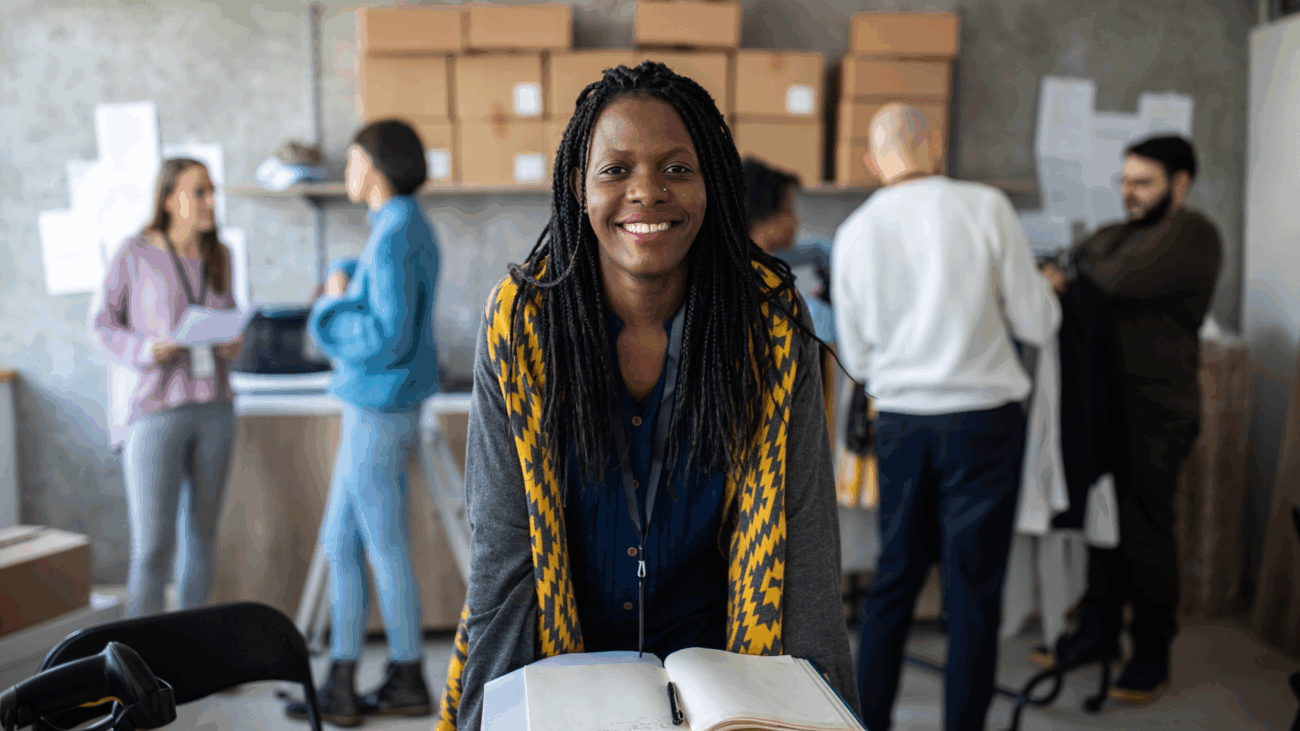 A smiling woman holds a book, symbolizing empowerment and support for black entrepreneurs in a vibrant marketplace.