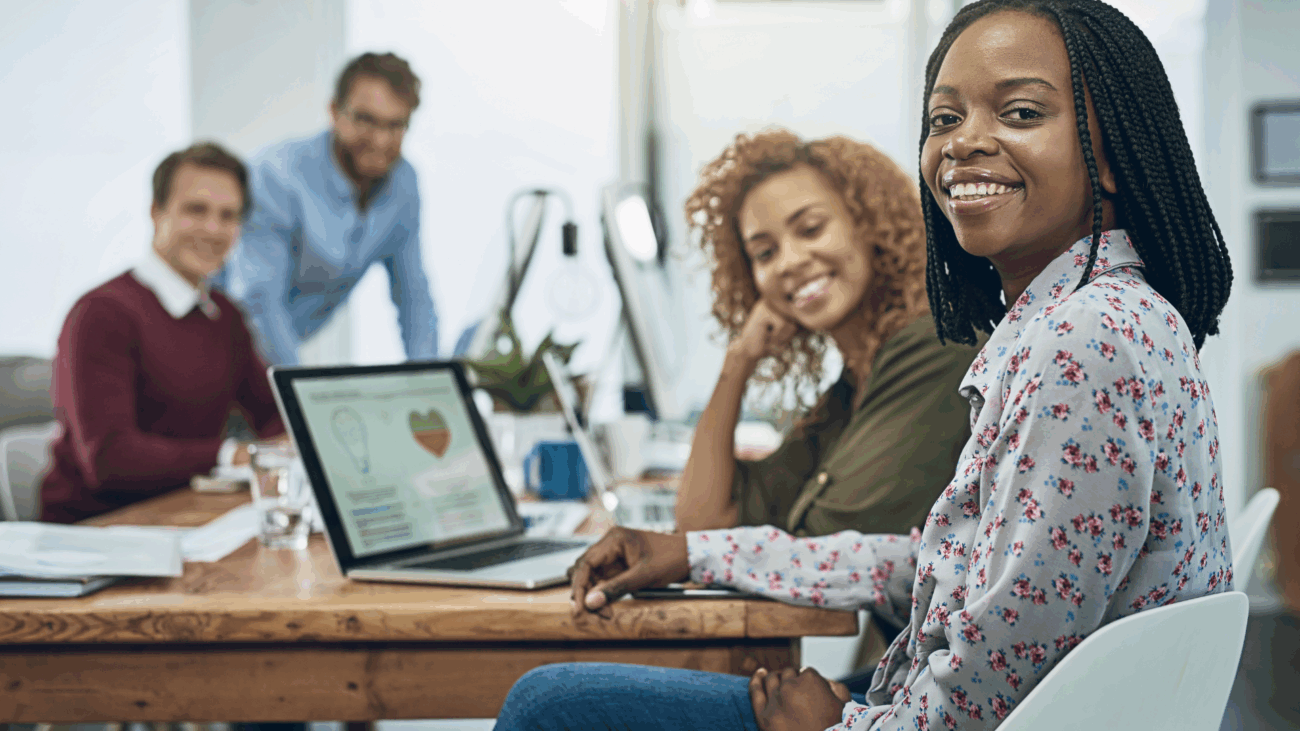 A smiling woman sits at a table with her laptop, embodying empowerment and community in a vibrant workspace.